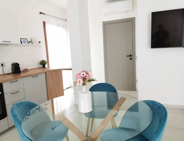 Zona pranzo moderna di Domus Luna con tavolo circolare in vetro, sedie di design in velluto azzurro e vista sulla cucina moderna open space.