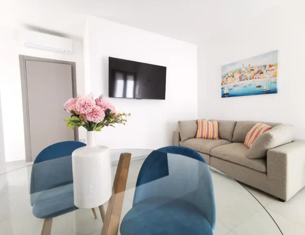 Soggiorno open space dell'appartamento Domus Luna con tavolo da pranzo, sedie di design azzurre e divano accogliente in ambiente luminoso.